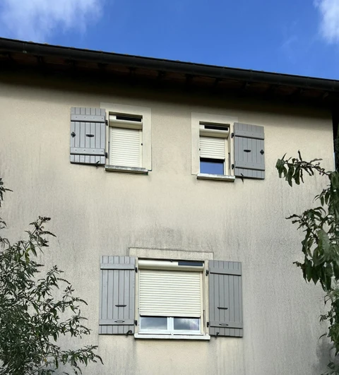 Façade de maison résidentielle avec trois fenêtres équipées de volets battants gris et stores enrouleurs blancs partiellement fermés